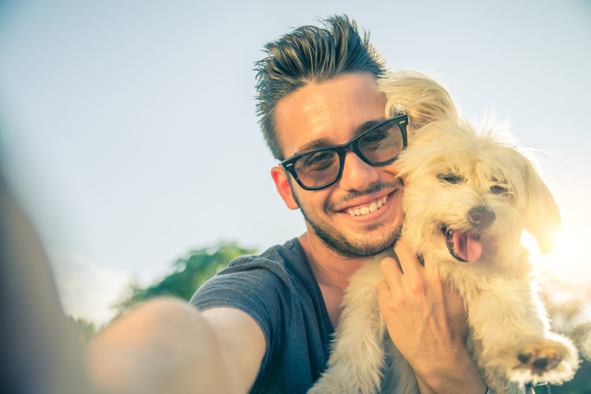 Man in black rimmed glasses holding up a small dog and taking a selfie with the sun shining behind them.